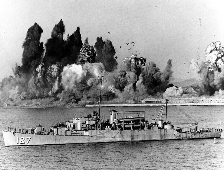 During the Evacuation of Hungnam, 24 December 1950, USS Begor (APD-127) stands offshore, ready to embark the last UN landing craft, as demolition charges wreck Hungnam's port facilities. (U.S. National Archives)